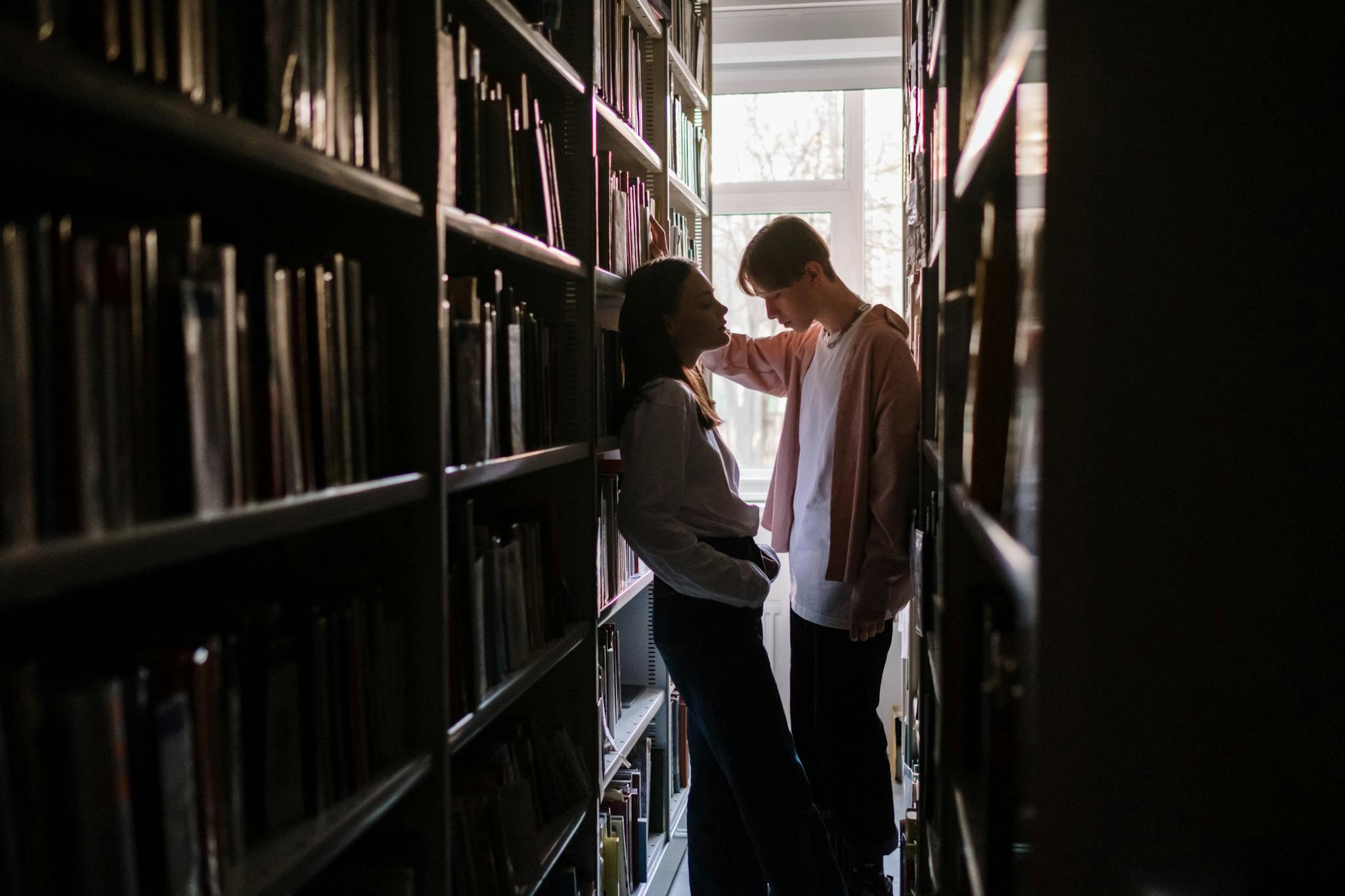 Two young adults having a discussion in a library aisle with natural light filtering in.